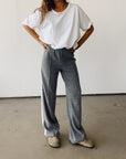 Indoors, a person wears Hey Gang's The Track Pant—high-waisted, gray, wide leg, tri-blend fleece—paired with a loose white tee and tan slippers. Their hands are on their hips; their head is not visible.