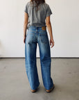 A woman stands indoors facing away, wearing Hey Gang's The Wide Slouch Jean in Harris wash with a gray t-shirt and brown shoes, set against a plain white wall and concrete floor.