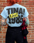 A person with red hair and colorful tattoos faces a brick wall, wearing the DayDreamer Tina Turner 1985 Reverse DF Tee, which features bold "TINA! 1985 Live!" text on the back. They’re holding a black and red scarf.