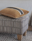 The Eastern Accents TAOS FAUX MOHAIR pillow, featuring two dark textured stripes and subtle beaded trim, sits on a patterned upholstered ottoman with wooden legs beside a gray textured surface and woven rug.