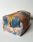 The Faire Dyed Toiletries Bag, made from 100% cotton with a yellow zipper and hand-dyed blue and peach tie-dye design, is displayed on a plain white surface.