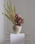 Tomar Vase by Accent Decor with tall green stems and purple spotted flowers on a sleek white pedestal.