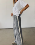 A person stands indoors, hands on hips, wearing Hey Gang’s The Track Pant—gray wide-leg pants with a white side stripe—and a loose white t-shirt against a plain, minimalistic background.