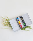 The Faire Travel Watercolor Paint Palette, featuring twelve vibrant pans, is open on a white surface with delicate white and yellow flowers and green leaves arranged beneath it.