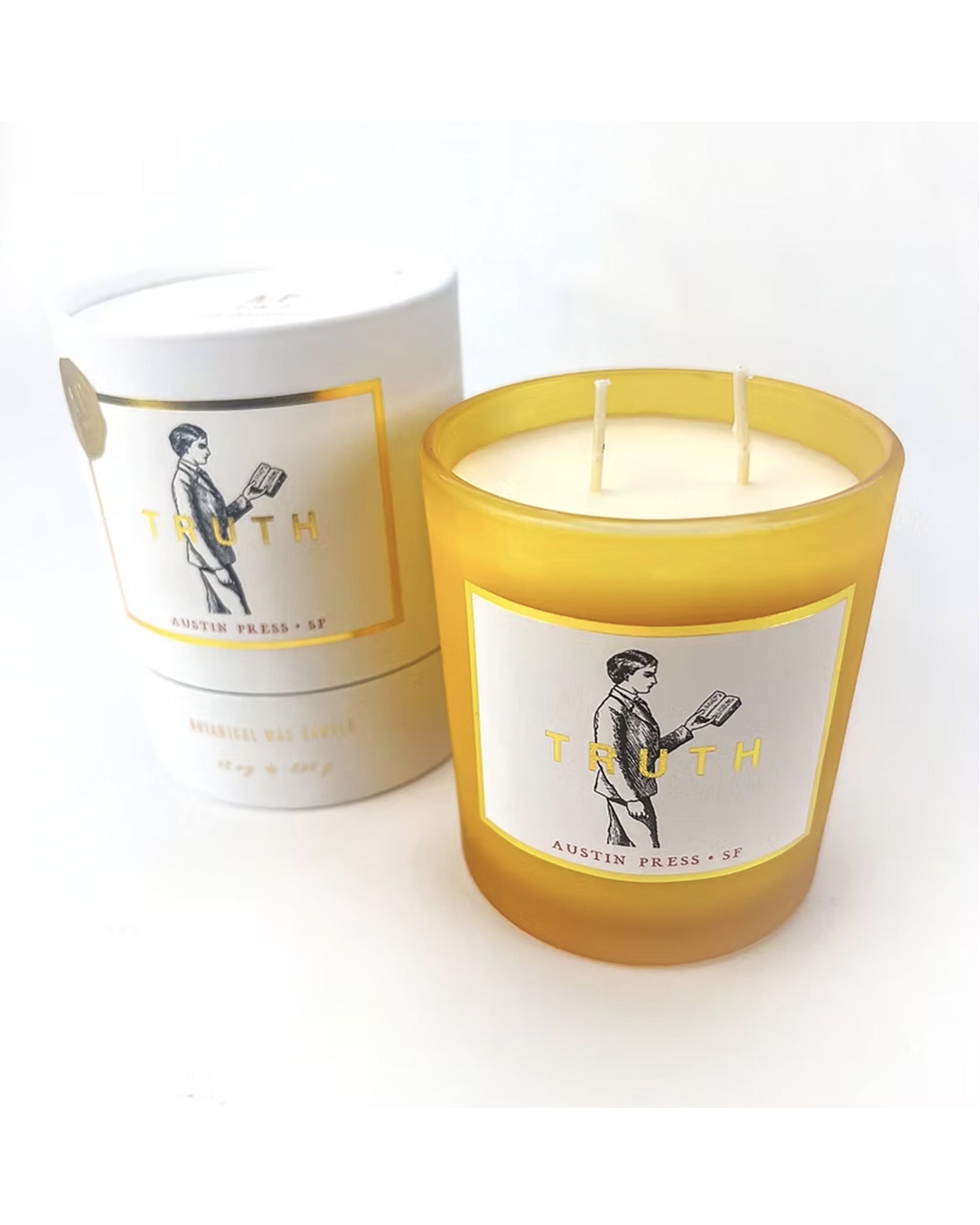 Yellow glass Truth Candle by Faire, featuring a person holding a book, comes with a matching white and yellow box. This two-wick candle is made using natural essential oils for a pure fragrance.