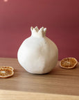 The Faire Turner Pomegranate, a white ceramic sculpture, rests on wood beside two dried orange slices. Set against a muted red background, it adds rustic charm to any home décor.
