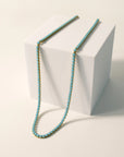 The Faire Turquoise Tennis Necklace features turquoise rhinestones on a hypoallergenic, 18K Gold PVD coated stainless steel chain, draped over a white cube and displayed against a light, neutral background.
