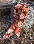 A spool of barbed wire rests outdoors amid dry grass, with the Faire Rustic Wild Rose Twilly Scarf—a polyester-silk blend featuring hand-drawn cream rose patterns—draped gracefully over it.
