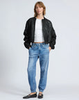 Wearing a black textured bomber jacket, white t-shirt, black flats, and Rag & Bone's Miramar Jogger—a blue denim-print jogger-style jean—a person stands confidently with hands in pockets against a plain backdrop.