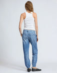 A person with light brown hair faces away, wearing a white tank top, blue high-rise Miramar Jogger jeans by Rag & Bone, and black ballet flats against a plain light gray background.