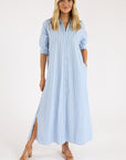 A woman poses against a plain background wearing the Xirena Boden Dress, a light blue, ankle-length cotton poplin shirtdress with elbow-length sleeves and side slits, styled with nude heeled sandals.