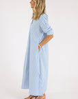A woman in profile wears the Xirena Boden Dress—a long, light blue cotton poplin shirtdress with vertical white stripes. She has wavy, shoulder-length blonde hair, nude heeled sandals, and her hands in the dress pockets.