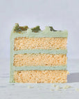 A slice of three-layer vanilla cake made with Wild Rye Cake Mix, featuring light green frosting between layers and on top, sits on a light surface with a few cake crumbs nearby—perfect for vanilla lovers.