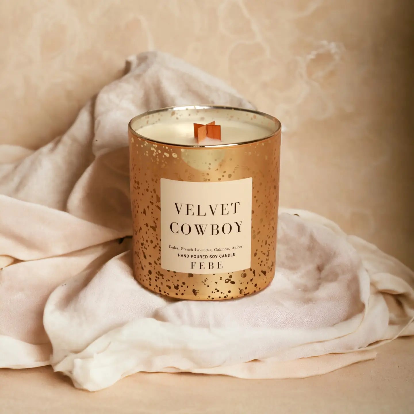 A scented candle labeled "Velvet Cowboy" by Faire, with a single wick, displayed on a soft, crumpled fabric against a marbled beige background. The FEBE candle's container has a speckled gold Arizona-style design.