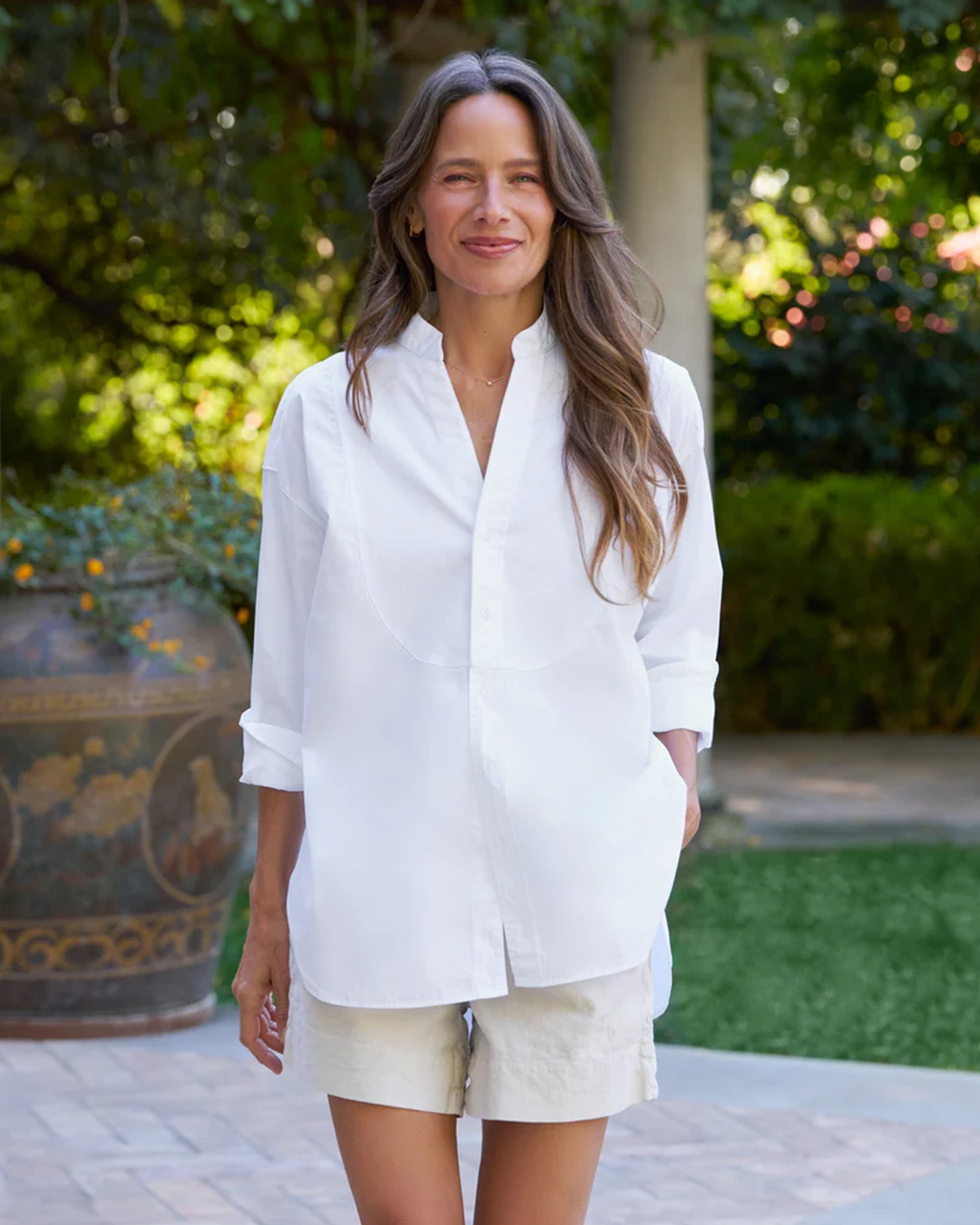 A woman with long brown hair smiles warmly outdoors, hands in her pockets, wearing the Frank & Eileen Victoria Collarless shirt and light-colored shorts, standing amid garden greenery and a large decorative vase.