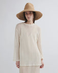 A person in the Janessa Leone Waverly Sand raffia straw hat with a shapeable brim wears a sheer, long-sleeved off-white top, standing against a plain light background and looking at the camera neutrally.
