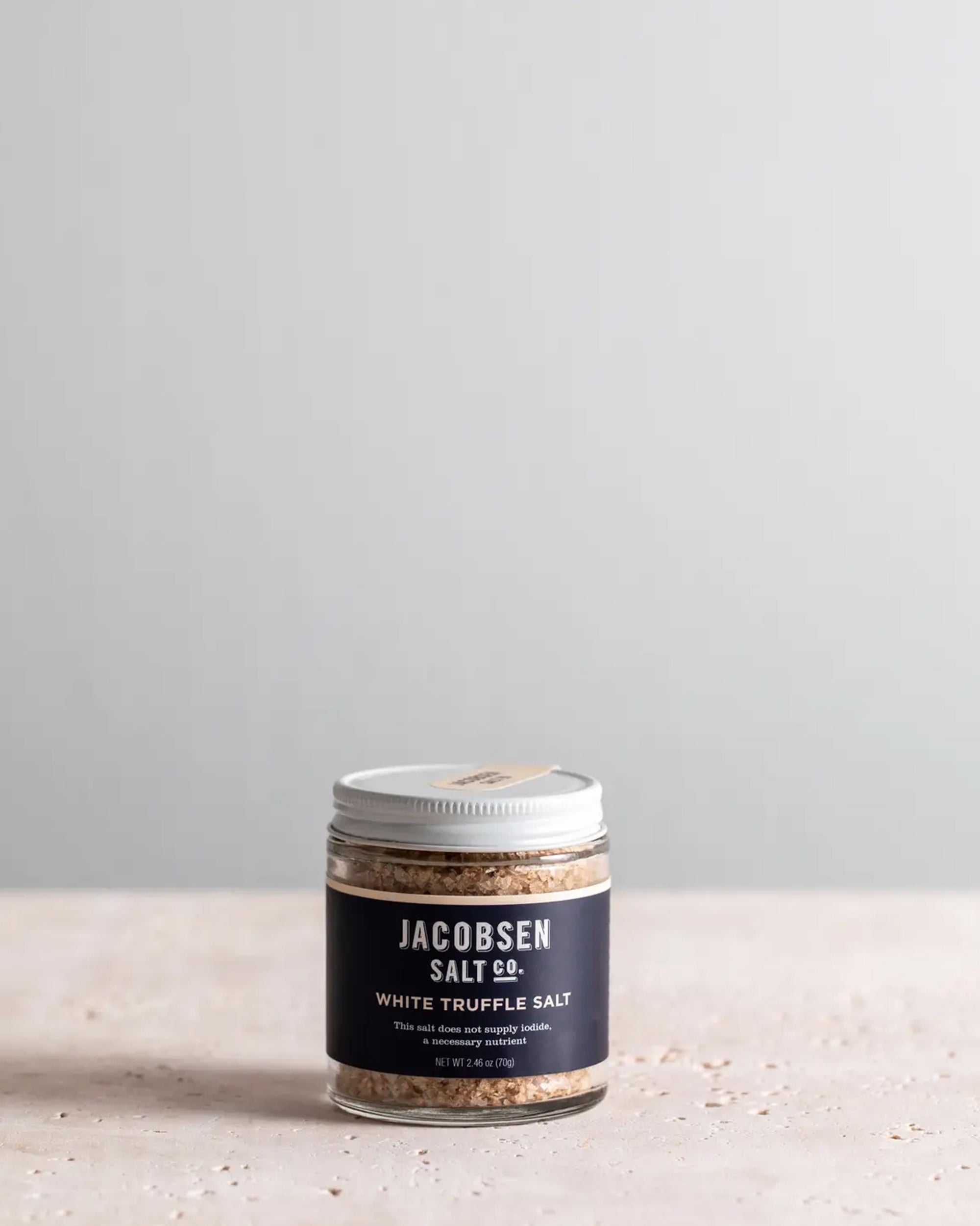 A jar of Faire White Truffle Salt with a silver lid rests on a light textured surface, set against a plain grey background. Its dark label highlights the premium salt infused with rich white truffle flavor.