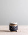 A jar of Faire White Truffle Salt with a silver lid rests on a light textured surface, set against a plain grey background. Its dark label highlights the premium salt infused with rich white truffle flavor.