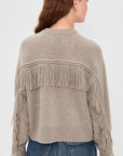 A person with long, wavy brown hair is seen from behind wearing the White + Warren Cashmere Luxe Fringe Crew in taupe, paired with blue jeans. The oversized sweater features fringe on the back and sleeves, set against a light-colored background.