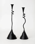The Faire Winding Taper Candle Holders feature black powder-finished aluminum with twisted, wavy stems and wide, uneven bases. Their unique, sculptural design adds a modern touch to any décor. Set of two shown against a plain white background.