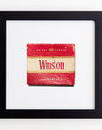 A framed picture of a Winston cigarette pack with a red top and bottom, and a cream-colored middle section. The pack features the words "Filter Tipped" and "Cigarettes" printed on it. Encased in clear acrylic, the frame is black with a white background. The product is called Art Square Blk Frame by Match South.
