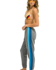 Woman in yellow sunglasses wears a rainbow bikini top and Aviator Nation 5 Stripe women's sweatpants, featuring blue side stripes, standing barefoot against a white background and looking to the side.