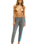 A woman stands barefoot against a white background, wearing a gold bikini top with red and black accents, yellow-tinted sunglasses, and Aviator Nation’s 5 Stripe Women’s Sweatpant with a blue side stripe.