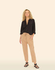 A person with curly blond hair confidently stands against a cream background, wearing a black button-up shirt and Xirena's beige Draper Pants with a drawstring waist. They sport black sandals and look directly at the camera.