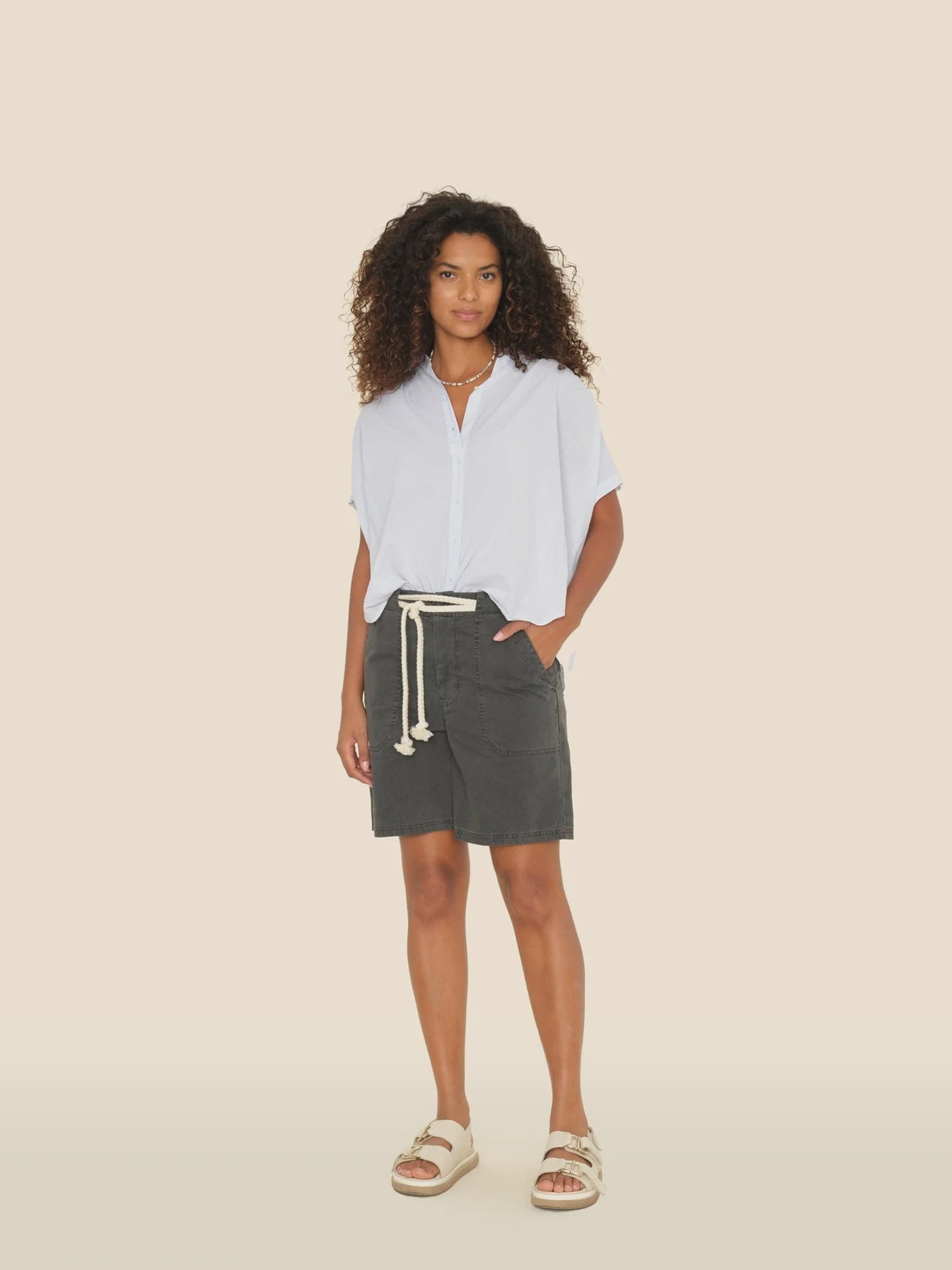 A person with curly hair wears Xirena's Toby Short, a light blue vintage washed cotton short-sleeve shirt, paired with a dark skirt featuring a rope belt and beige sandals, standing against a plain background.