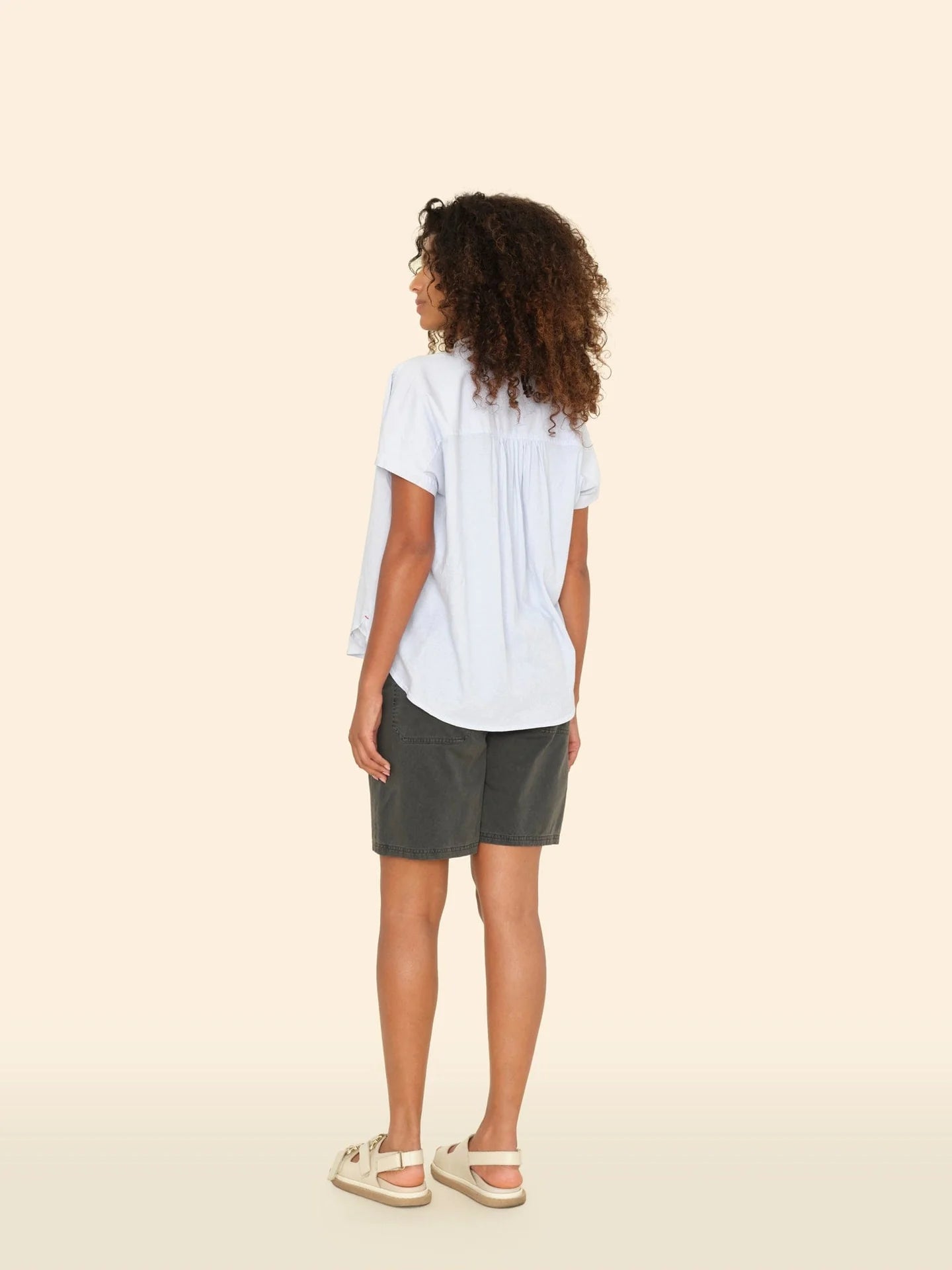 A curly-haired woman in a light blue blouse and Xirena's Toby Short stands facing away against a beige backdrop, wearing white slip-on shoes and exuding effortless Los Angeles style.