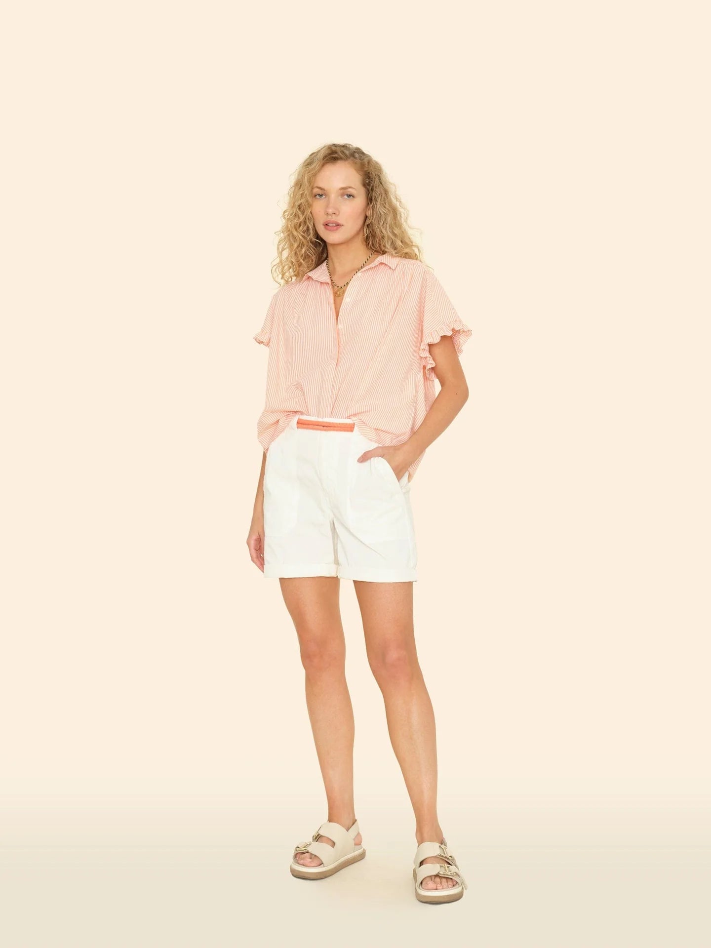 A curly-haired individual is wearing a pink short-sleeve button-up shirt with Xirena's Toby Short in white. Beige sandals complete the relaxed look, all set against a plain light background, ideal for casual workwear days.
