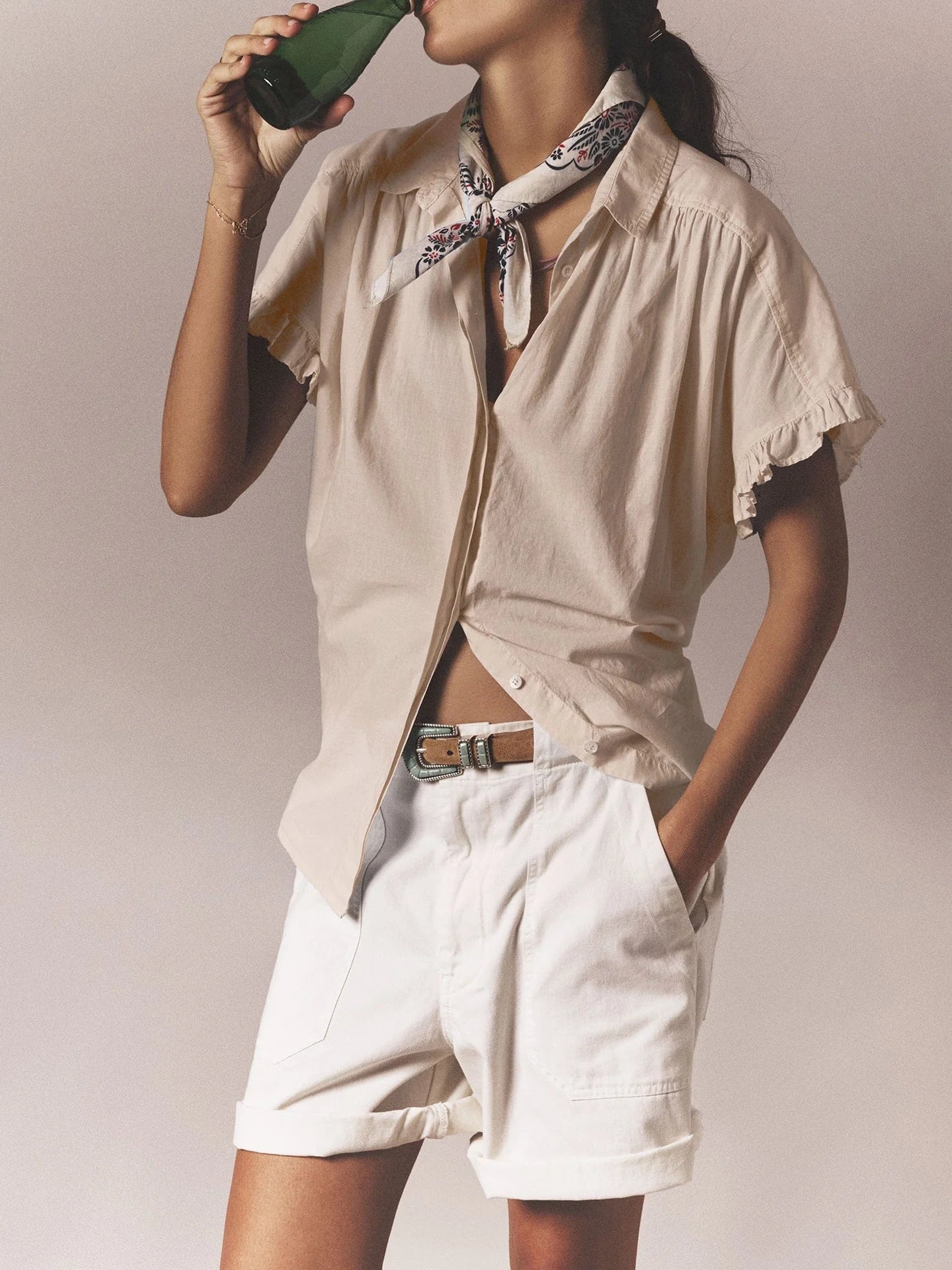 A person in a light beige short-sleeve blouse and Xirena's Toby Short with patch pockets drinks from a dark bottle. A scarf is around their neck, and their left hand rests in the pocket against a backdrop reminiscent of vintage washed cotton twill.