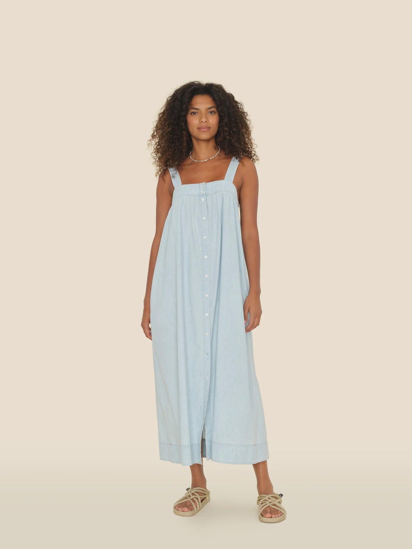A woman with curly hair wears the Xirena Orly Dress, a light blue button-front cotton chambray midi sundress with adjustable straps, and beige sandals. She stands against a plain light beige background, blending practicality and elegance.