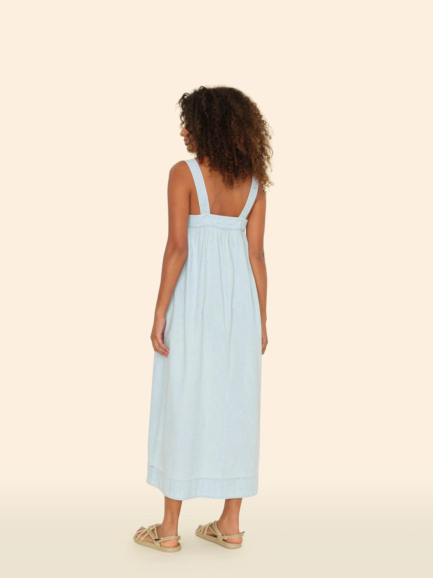 A person with curly hair stands facing away in the Orly Dress by Xirena, a sleeveless, ankle-length light blue cotton chambray sundress with wide adjustable straps. They pair it perfectly with beige sandals against a plain light beige background.