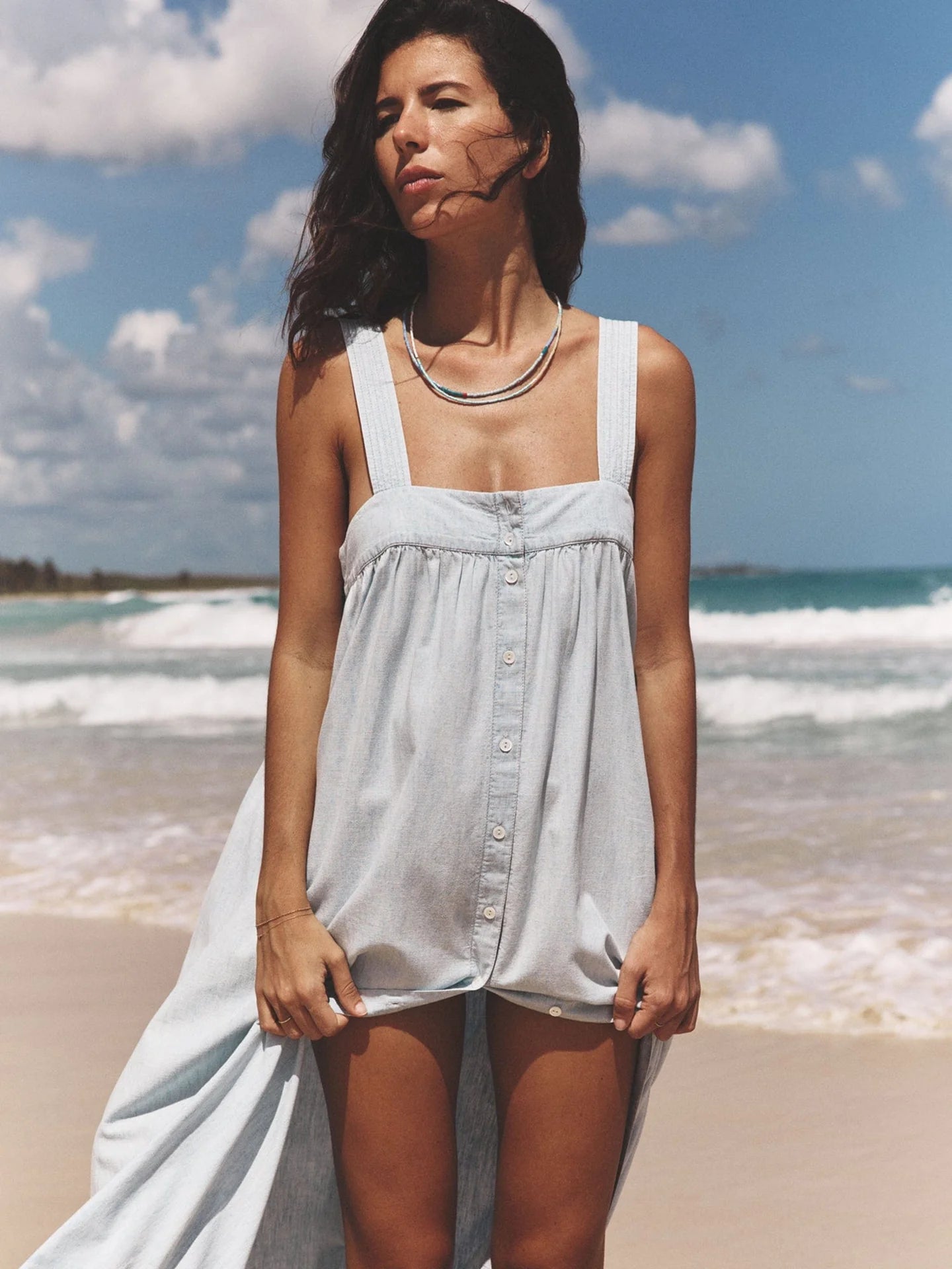 A woman stands on a beach wearing the Orly Dress by Xirena, a cotton chambray sundress with buttons and adjustable straps. Her necklace catches the sunlight as she gazes into the distance, while the serene ocean and cloudy sky form her backdrop.