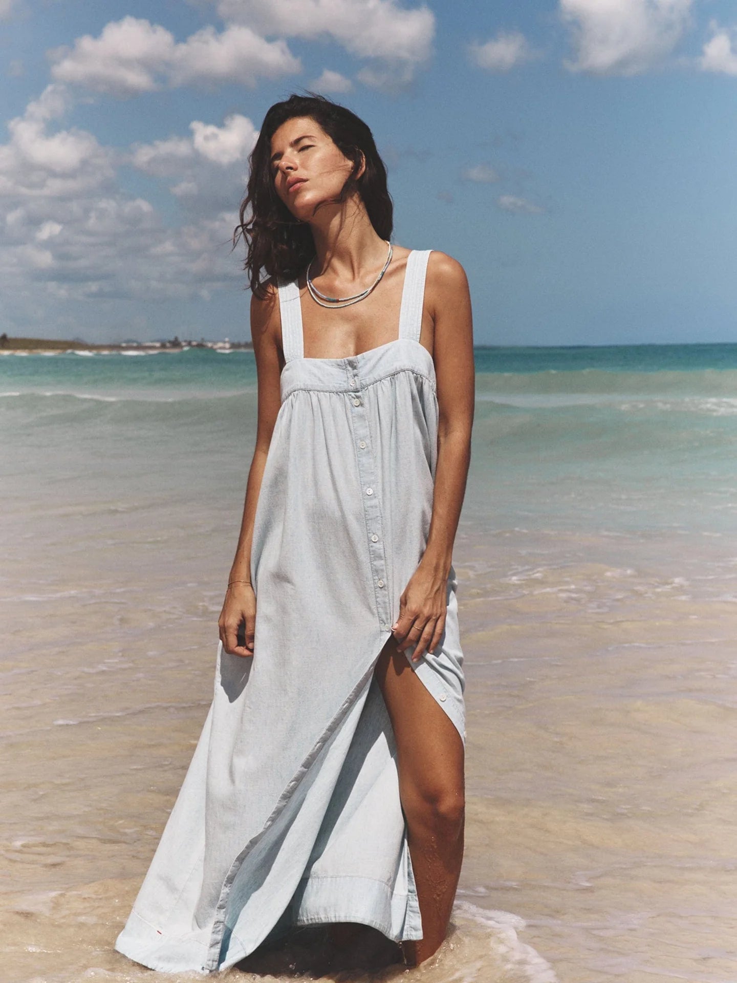 A woman wearing the Orly Dress by Xirena, a light blue cotton chambray sundress with adjustable straps, stands on a sandy beach gazing upward with a relaxed expression. The sky is partly cloudy, and the vibrant blue ocean waves gently lap the shore.