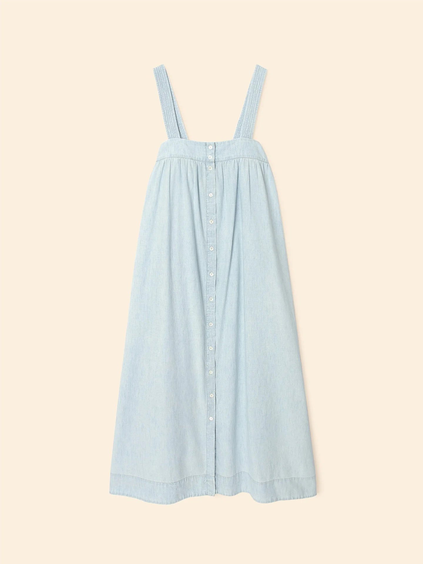 The Xirena Orly Dress is a light blue cotton chambray sundress with wide shoulder straps and a button-down front. It features a gathered waist and flowing, knee-length skirt on a plain beige background.