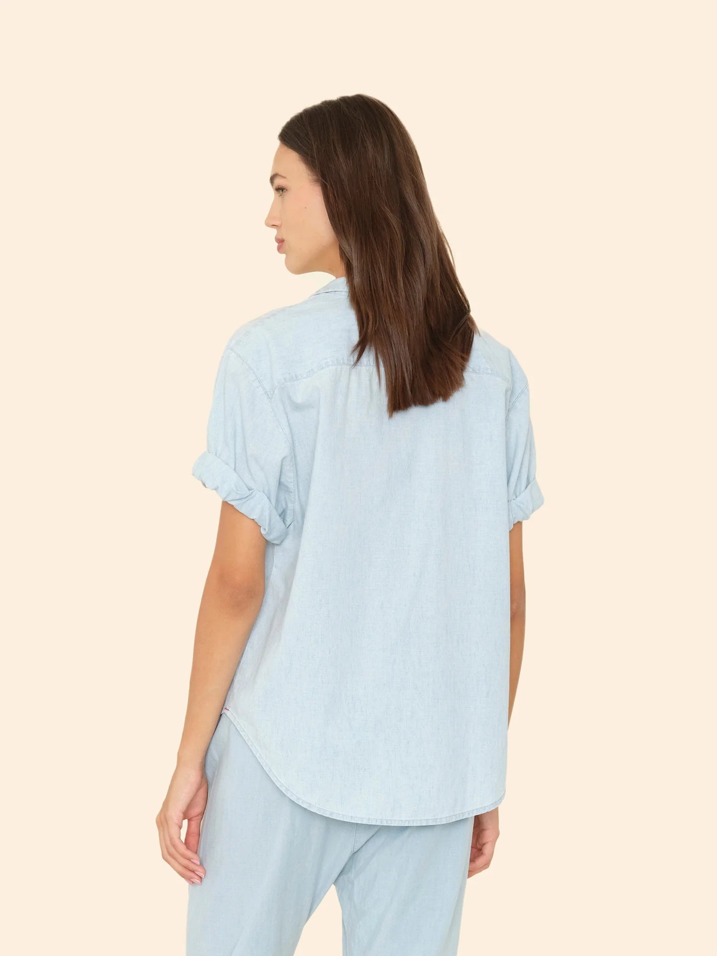 A woman with long brown hair wears the Xirena Channing Shirt S25 in light blue chambray with matching pants, standing sideways against a beige background.