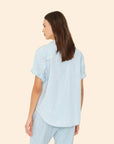 A woman with long brown hair, seen from behind, wears the light blue cotton chambray Xirena Channing Shirt S25 and matching pants against a plain beige background.