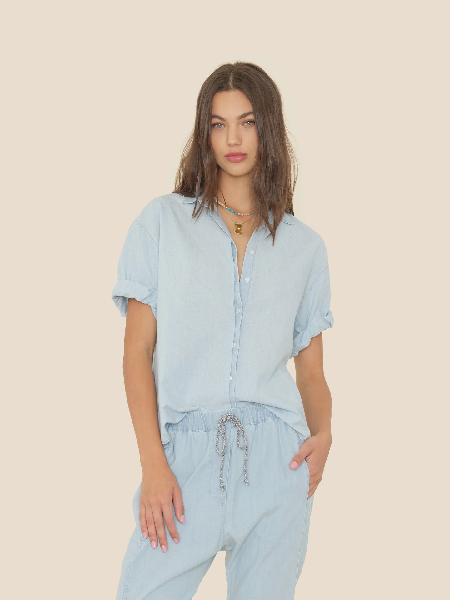 A woman with long brown hair wears the Xirena Channing Shirt S25 in soft cotton chambray with matching drawstring pants, standing against a beige background. She accessorizes with layered necklaces and has one hand in her pocket.