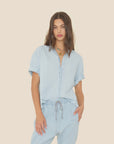 A woman with long brown hair wears the Xirena Channing Shirt S25 in soft cotton chambray with matching drawstring pants, standing against a beige background. She accessorizes with layered necklaces and has one hand in her pocket.