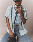 A person wears the Xirena Channing Shirt S25 over a blue top, light blue jeans, a patterned scarf, cap, and bracelets, standing stylishly against a plain background.
