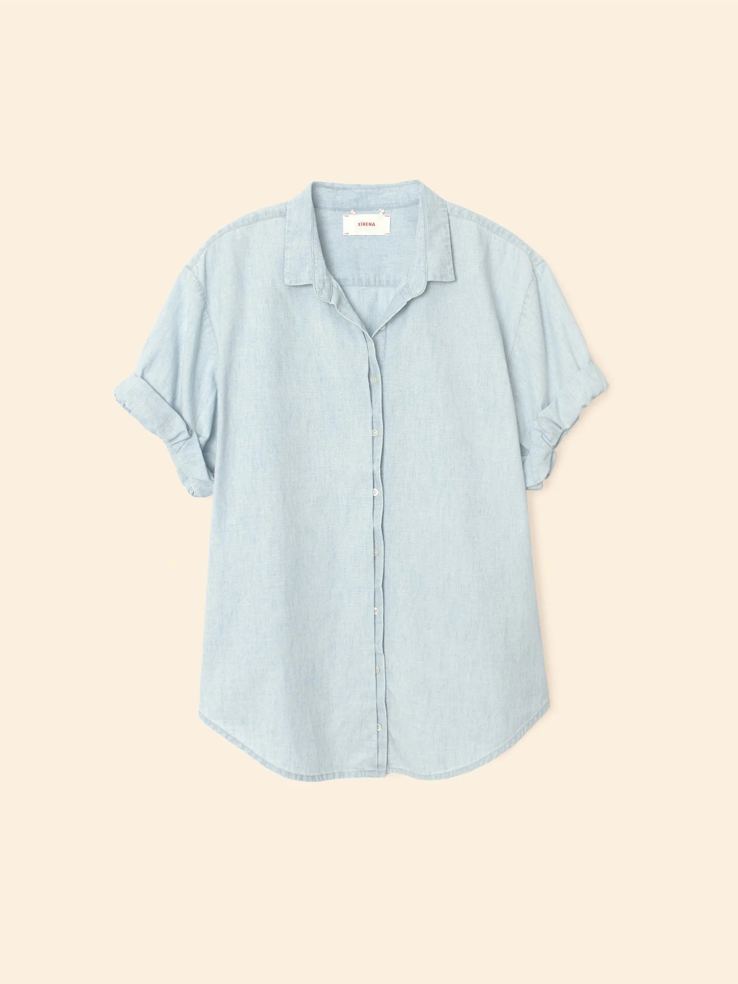 The Xirena Channing Shirt S25 in light blue cotton chambray features short sleeves, rolled cuffs, a small collar, and a relaxed fit, shown against a plain beige background.