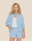 A woman with curly blonde hair wears Xirena's Jordy Shirt, featuring blue and white stripes, over a white top with matching shorts. Her left hand rests in her pocket against a plain beige background.