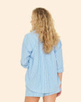 A person with long curly blonde hair, turned away, wears the Jordy Shirt by Xirena with matching shorts in light blue and white pencil stripes against a solid beige background.