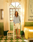 A woman stands in a sunlit room with checkered floor tiles, wearing the Jordy Shirt from Xirena and white shorts. She leans on a white chair by a window with curtains. The room features green walls, a yellow bedspread, and a vintage poster.