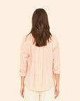 A person with long brown hair facing away is wearing the Jordy Shirt by Xirena, a light pink, button-front shirt with pencil stripes. It pairs perfectly with light-colored pants against a beige background.