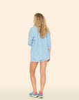 A person with curly blonde hair, facing away, is wearing the Carlyn Short by Xirena in a crisp cotton light blue set, including a button-up shirt and matching shorts. They're paired with colorful sneakers against a plain beige backdrop, reminiscent of sunlit Los Angeles afternoons.