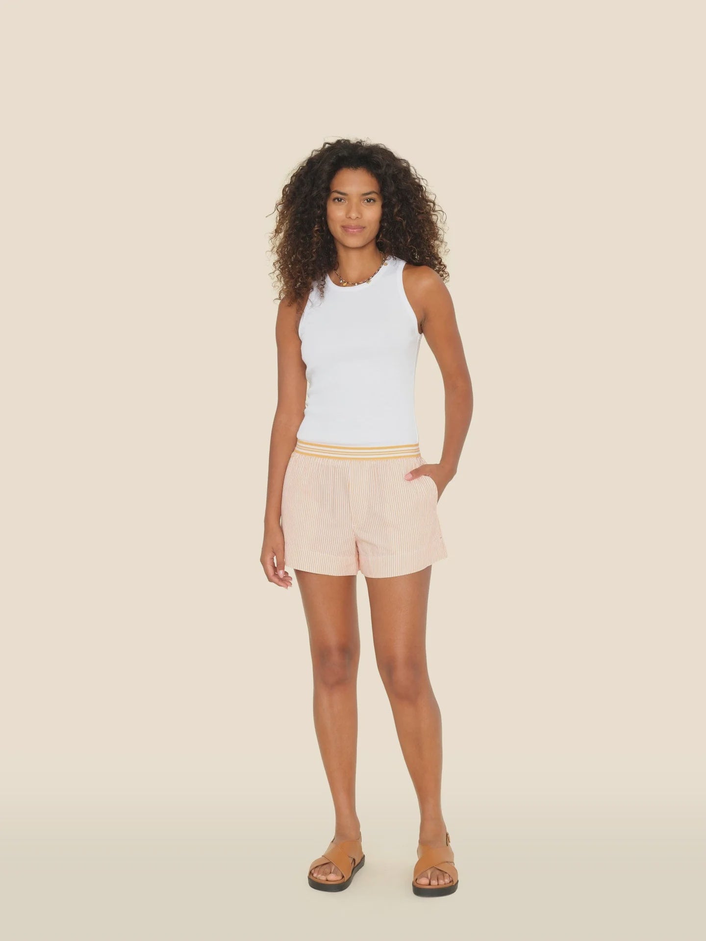 A person with curly hair poses against a plain background in a white sleeveless top and Xirena's Carlyn Shorts, highlighted by a yellow elasticized waistband. Brown sandals complete the look, with one hand in their pocket and the other by their side.