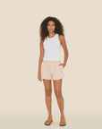 A person with curly hair poses against a plain background in a white sleeveless top and Xirena's Carlyn Shorts, highlighted by a yellow elasticized waistband. Brown sandals complete the look, with one hand in their pocket and the other by their side.